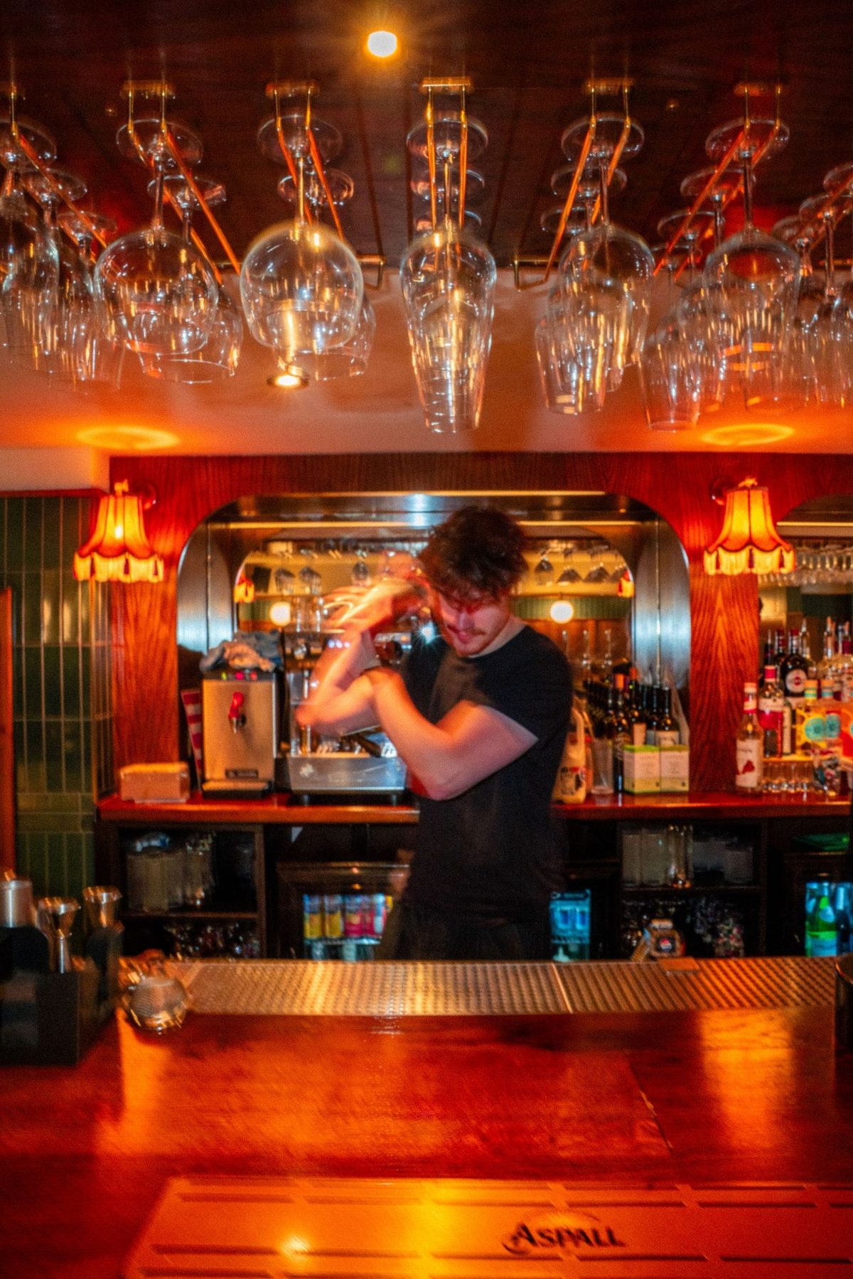 A bartender shaking a cocktail behind the bar, glasses hanging overhead